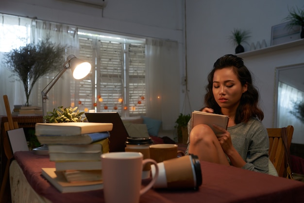 A student sitting at a desk in a dorm room, studying with books and a laptop. The scene is brightly lit, suggesting diligence and focus.
